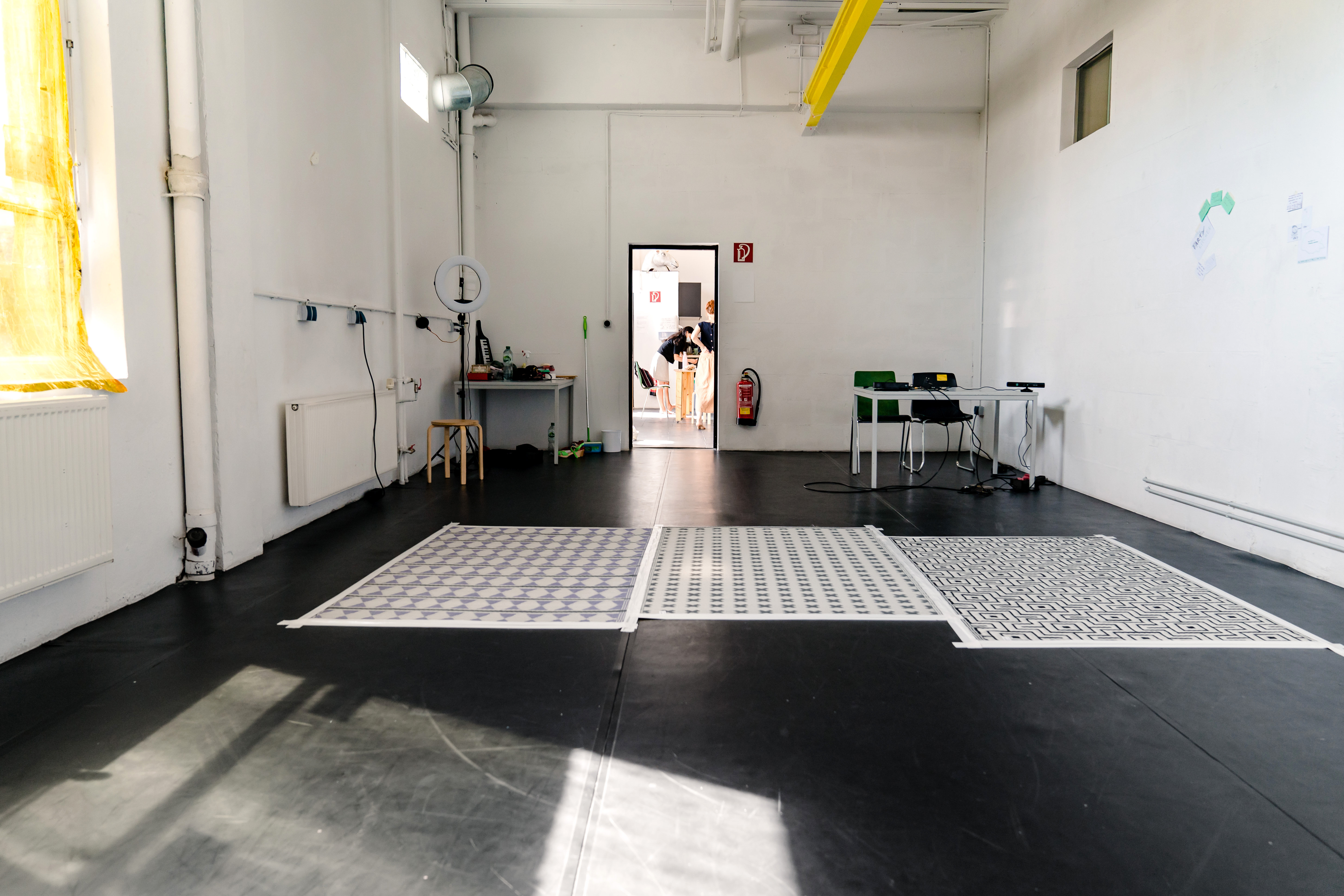 Kleines Studio Ein Raum mit weißen Wänden, einem Fenster, drei aufgeklebten Teppichen auf dem dunklen Boden. Durch die geöffnete Tür sieht man zwei Personen.